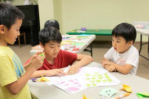 Eight-year-old Derek (left) creates an origami frog with library volunteer, Kevin (center), and four-year-old Alex (right). Photos by Laura Drinan